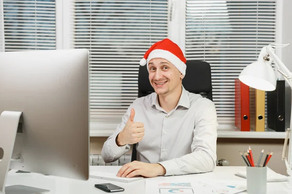 Schöner lächelnder Geschäftsmann in Hemd und roter Weihnachtsmütze am Schreibtisch sitzend, Daumen hoch, Arbeit am Computer mit modernem Monitor, mit Dokumenten im hellen Büro, Blick in die Kamera. — Stockfoto