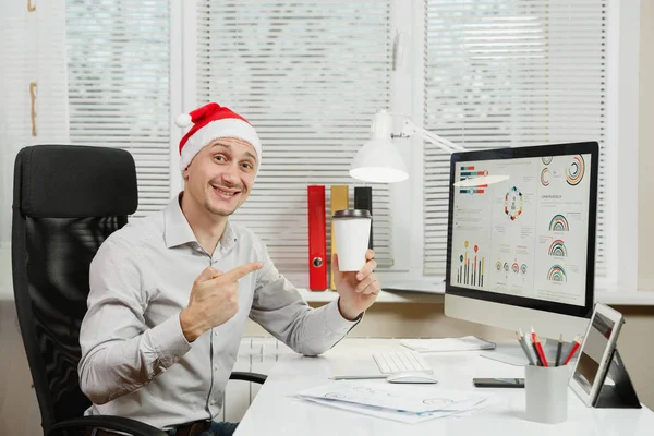 Hombre de negocios guapo sonriente con camisa y sombrero rojo de Navidad sentado en el escritorio con taza de té o café, trabajando en la computadora con monitor moderno, con documentos en la oficina de luz, mirando a la cámara . — Foto de Stock