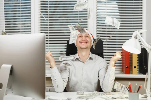 Sonriendo feliz hombre de negocios feliz en camisa, sombrero rojo de Navidad sentado en el escritorio con mucho dinero en efectivo, trabajando en el nuevo año en el ordenador con monitor moderno, lámpara, carpetas, documentos en la oficina de luz . —  Fotos de Stock