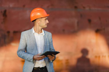 Yakışıklı tıraşsız başarılı iş adamı gri takım elbise, tablet, liman kargo paslı gemi arka plan ayakta tutan koruyucu inşaat turuncu kask. Erkek ile güneşli günde becerikli alet