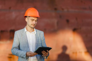 Yakışıklı tıraşsız başarılı iş adamı gri takım elbise, tablet, liman kargo paslı gemi arka plan ayakta tutan koruyucu inşaat turuncu kask. Erkek ile güneşli günde becerikli alet