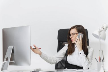 Takım elbise ve masa başında oturan gözlük güzel ciddi iş kadını ışık Office belgeleri ile çağdaş bilgisayarda çalışan cep telefonu beyaz arka plan üzerinde sorunlarını çözme konuşurken