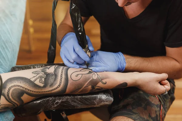 Close up of professional tattooer artist doing picture on hand of man by machine black ink from a jar. Tattoo art on body. Equipment for making tattoo art. Master makes tattooed in light studio.