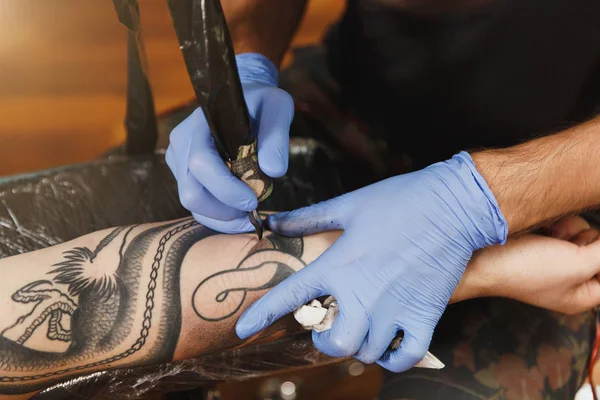 Profesyonel tattooer sanatçı tarafından makine siyah mürekkep bir kavanoz gelen adamın elini resim yapıyor kapatın. Vücudunda dövme sanatı. Dövme sanat yapmak için ekipman. Ana ışık studio dövmeli yapar.