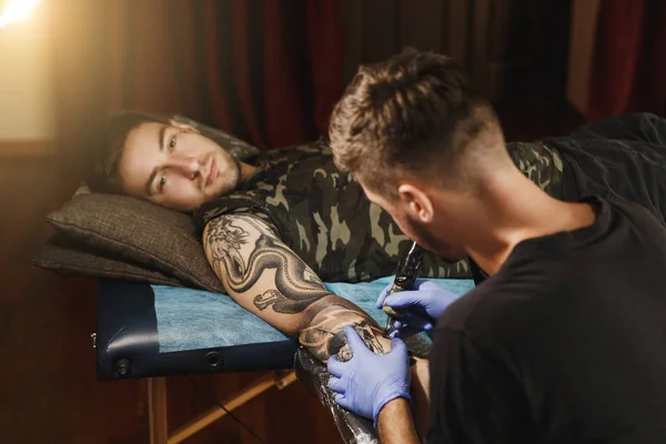 Профессиональный татуировщик рисует на руке человека черными чернилами из банки. Татуировка на теле. Оборудование для изготовления татуировок. Мастер делает татуировки в светлой студии . — стоковое фото