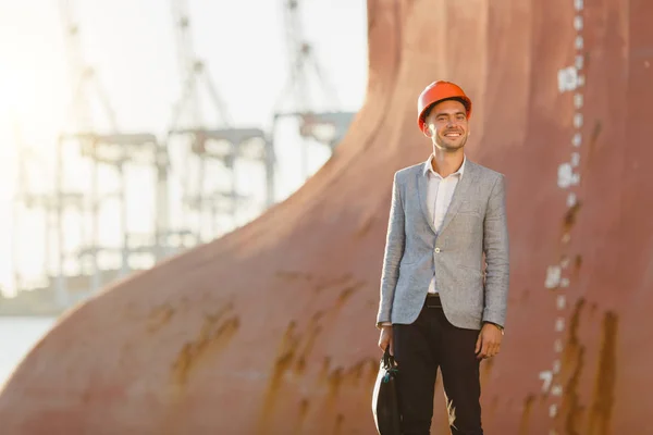 Handsome young unshaven successful business man in gray suit and protective construction orange helmet holding case, standing in sea port against a cargo rusty ship with water line background