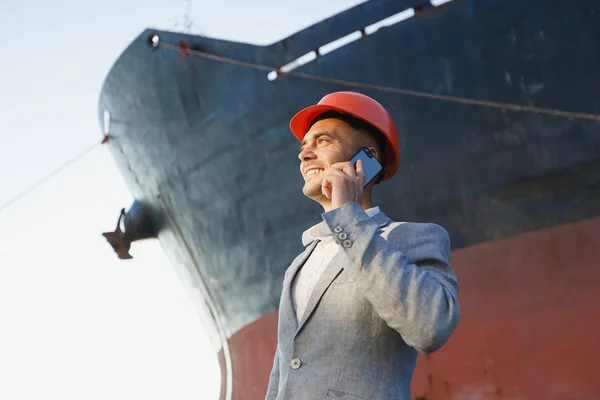 Handsome young unshaven successful business man in gray suit and protective construction orange helmet talking on mobile phone, standing in sea port against cargo rusty ship background. Bottom view
