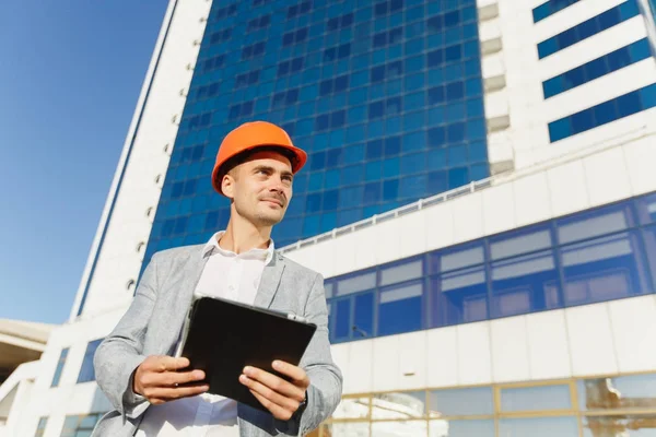 회색 양복과 흰색 파란색에서 건물의 배경에 태블릿 보호 건설 오렌지 헬멧 성공적인 비즈니스 사람. 오피스 빌딩에 노동자 보안에 대 한 헬멧에서 엔지니어. — 스톡 사진