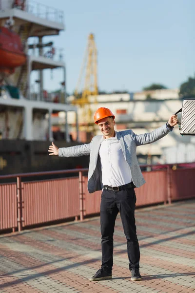 Handsome young unshaven successful business man in gray suit and protective construction orange helmet holding case, spreading hands, walking in sea port against cargo ship and crane background.