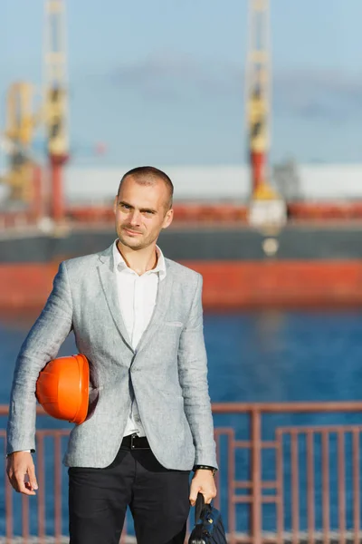 Handsome young unshaven successful business man in gray suit and protective construction orange helmet holding case, walking in sea port against cargo ship and crane background in summer time.