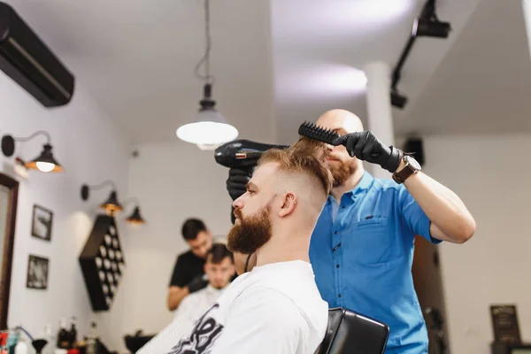 Мужчина профессиональный парикмахер, обслуживающий клиента, сушка волос с феном. Рыжий красивый грубый стильный молодой человек с густой бородой и короткими волосами, стригущийся модно. Светло-белый парикмахерская . — стоковое фото