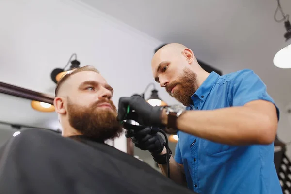 Cabeleireiro profissional masculino servindo cliente por clipper. Gengibre bonito jovem elegante brutal com barba grande grossa e cabelo curto ficando moderno corte de cabelo em capa preta. Sala de barbeiro branco claro — Fotografia de Stock