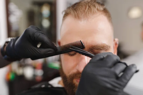 Male professional hairdresser serving client, shearing eyebrows with scissors. Ginger stylish young man with thick big beard and short hair getting trendy haircut in black cape. Light barber shop room