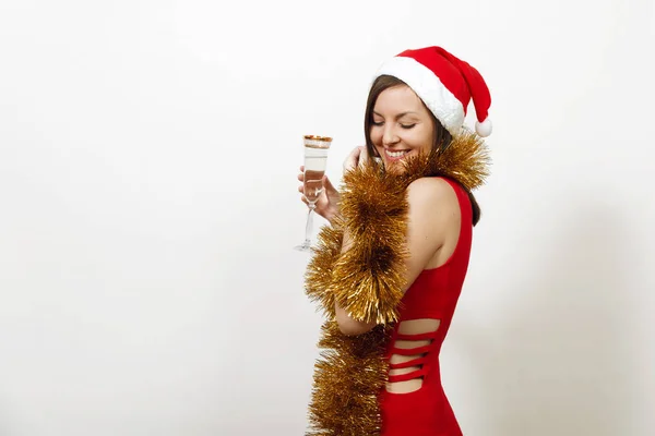 Bonito caucasiano sorrindo jovem mulher feliz em vestido vermelho, chapéu de Natal e guirlanda de ouropel de pé com copo de champanhe no fundo branco. Santa menina isolada. Conceito de férias de Ano Novo — Fotografia de Stock