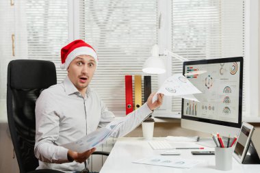 Pencere arka plan üzerinde ışık Office belgeleri ile modern monitör bilgisayarda çalışan yakışıklı şok ve çapraşık iş adamı masa başında oturan gömlek ve kırmızı Noel şapkalı. Yeni yıl.