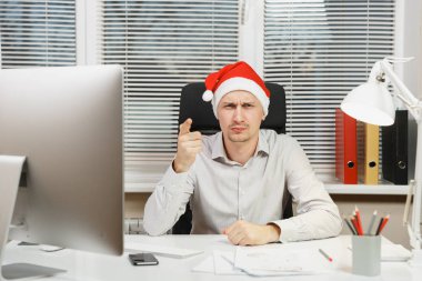 Masa başında oturan gömlek ve kırmızı Noel şapkalı yakışıklı üzgün ve kızgın iş adamı bilgisayar, yeni yılda modern monitör ile pencere arka plan üzerinde ışık Office belgeleriyle çalışma.