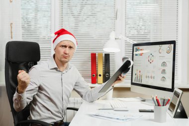 Pencere arka plan üzerinde ışık Office belgeleri ile modern monitör bilgisayarda çalışan yakışıklı şok ve çapraşık iş adamı masa başında oturan gömlek ve kırmızı Noel şapkalı. Yeni yıl.