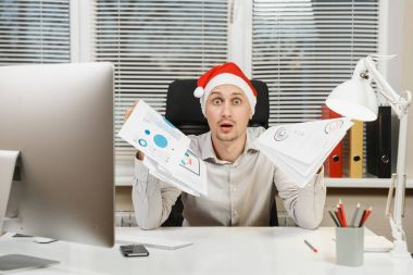 Yeni yıl bilgisayarda çalışan modern monitör, lamba, belge penceresi arka plan üzerinde ışık Office ile yakışıklı şok ve çapraşık iş adamı masa başında oturan gömlek, kırmızı Noel şapkalı.