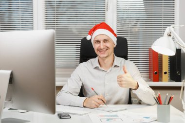 Başparmak gösterilmesini, bilgisayar ışık Office belgeleri ile modern İzleyicisi ile çalışma, kameraya bakarak yakışıklı gülümseyen iş adamı masa başında oturan gömlek ve kırmızı Noel şapkalı.