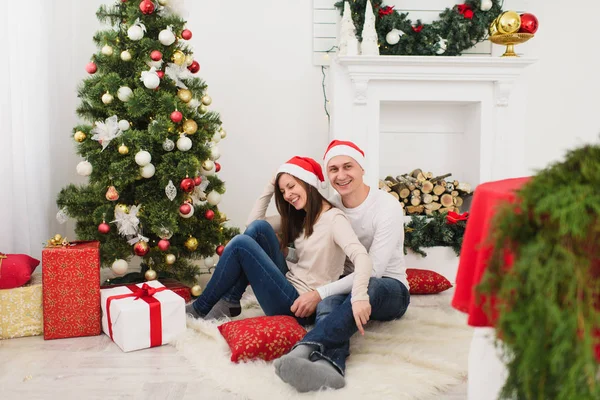 Gelukkig vrolijke grappige jonge verliefde paar in rode hoed zitten in lichte kamer thuis met ingerichte nieuwe jaar boom cadeau vakken en op de achtergrond van de kroon van Kerstmis. Familie, concept vakantie 2018. — Stockfoto