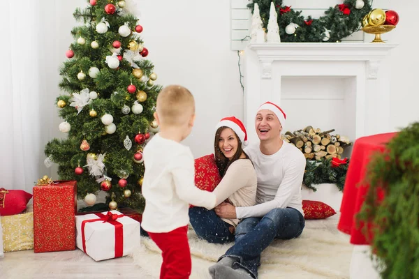 Happy young cheerful parents with cute little son in red hat. Child boy in light room at home with decorated New Year tree and gift boxes. Christmas good mood. Family, love and holiday 2018 concept. — Stock Photo, Image