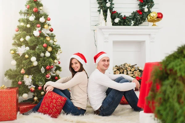 Gelukkig vrolijke grappige jonge verliefde paar in rode hoed zitten in lichte kamer thuis met ingerichte nieuwe jaar boom cadeau vakken en op de achtergrond van de kroon van Kerstmis. Familie, concept vakantie 2018. — Stockfoto