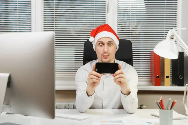 Guapo sonriente hombre de negocios divertido en camisa, sombrero rojo de Navidad sentado en el escritorio, haciendo selfie en el teléfono móvil, trabajando en el nuevo año en la computadora con monitor moderno, lámpara, documentos en la oficina de luz . — Foto de Stock