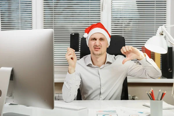 Un om de afaceri trist și supărat în cămașă și pălărie roșie de Crăciun stând la birou cu telefonul mobil, arătând degetul mare în jos, lucrând în noul an la calculator cu monitor modern, cu documente în birou luminos — Fotografie, imagine de stoc