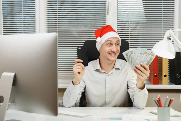 Sorrindo feliz homem de negócios feliz na camisa, chapéu de Natal vermelho sentado na mesa com telefone celular e muito dinheiro em dinheiro, trabalhando no ano novo no computador com monitor moderno, documentos no escritório leve . — Fotografia de Stock