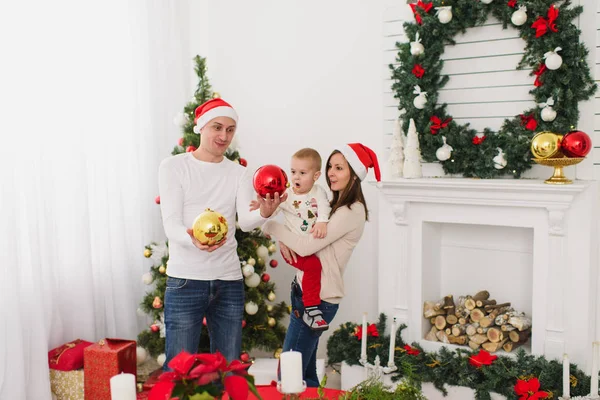 Szczęśliwy wesoły rodziców z cute synka. Chłopiec dziecko duży czerwony drzewo zabawka piłka stojący w urządzony nowy rok jasny pokój w domu na tle Boże Narodzenie wieniec. Rodzina, wakacje 2018 koncepcja. — Zdjęcie stockowe