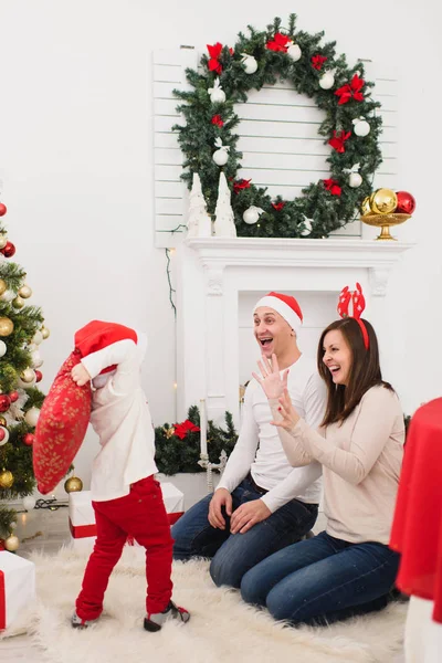 Happy young cheerful parents fighting pillows with cute little son. Child boy in red hat in light room at home with decorated New Year tree. Christmas good mood. Family, love and holiday 2018 concept. — Stock Photo, Image