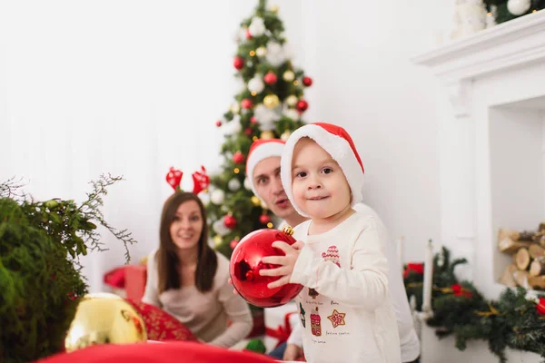 Padres jóvenes felices con lindo hijo en sombrero. Niño con gran bola de juguete de árbol rojo de pie en la sala de luz decorada de Año Nuevo en casa. Navidad buen humor. Familia, amor y vacaciones 2018 concepto . — Foto de Stock