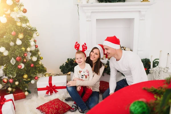 Pais alegres felizes com o filhinho bonito. Menino em chapéu vermelho sentado na sala de luz em casa com árvore de Ano Novo decorada e caixas de presente. Bom humor de Natal. Conceito de família, amor e férias 2018 . — Fotografia de Stock