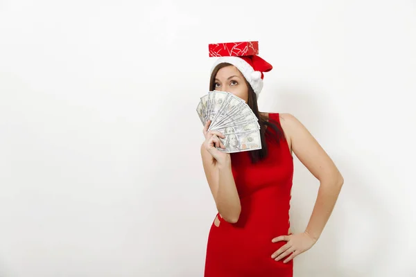 Krásná mladá šťastná žena v červených šatech a vánoční čepice drží dárkové krabičky skrývá tvář za peníze bankovky na bílém pozadí. Santa dívka s hotovostí, samostatný. Nový rok dovolená 2018 concept — Stock fotografie