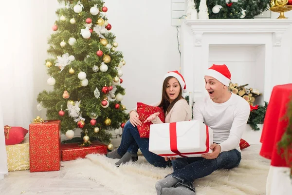 Feliz alegre divertida pareja joven enamorada en sombrero rojo sentado en la sala de luz en casa con árbol de Año Nuevo decorado y cajas de regalo en el fondo de la corona de Navidad. Familia, vacaciones 2018 concepto . — Foto de Stock