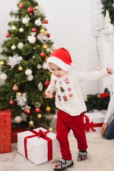 Happy cheerful cute little child boy in red hat and pants, running in light room at home with decorated New Year tree and gift boxes. Christmas good mood. Family, love and holiday 2018 concept. — Stock Photo, Image