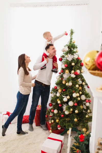 Happy young cheerful parents with cute little son. Child boy on daddy shoulders decorating New Year tree with toy ball in light room at home. Christmas good mood. Family, love and holiday 2018 concept — Stock Photo, Image