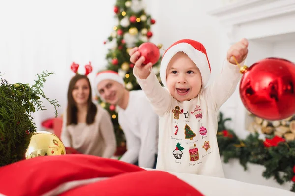 Padres jóvenes felices con lindo hijo en sombrero. Niño con gran bola de juguete de árbol rojo de pie en la sala de luz decorada de Año Nuevo en casa. Navidad buen humor. Familia, amor y vacaciones 2018 concepto . — Foto de Stock