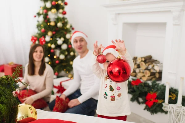 Padres jóvenes felices con lindo hijo en sombrero. Niño con gran bola de juguete de árbol rojo de pie en la sala de luz decorada de Año Nuevo en casa. Navidad buen humor. Familia, amor y vacaciones 2018 concepto . — Foto de Stock