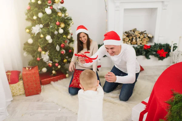 Feliz joven padres alegres con lindo hijo pequeño en sombrero rojo. Niño en la sala de luz en casa con árbol de Año Nuevo decorado y cajas de regalo. Navidad buen humor. Familia, amor y vacaciones 2018 concepto . — Foto de Stock