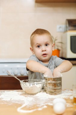 Küçük çocuk çocuk anne Noel ışık mutfakta zencefilli bisküvi pişirmek için yardımcı olur. Gri T-shirt hafta sonu sabah evde masada demir elek ve un ile 2-3 yıl içinde mutlu sarışın çocuk.
