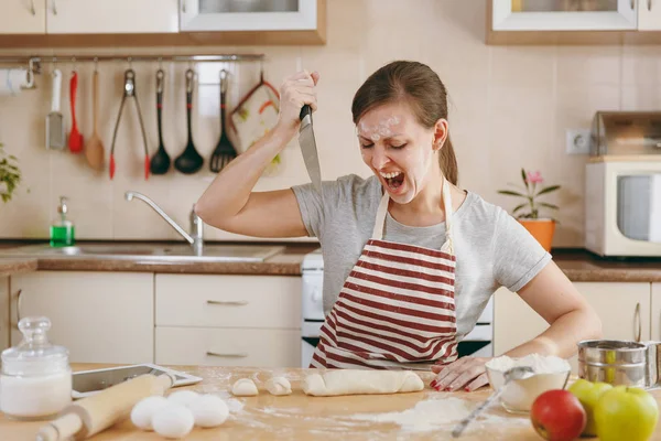 Młoda zła i agresywna kobieta siedzi przy stole i atakowanie ciasto z nożem w kuchni. Gotowanie w domu. Przygotowanie żywności. — Zdjęcie stockowe