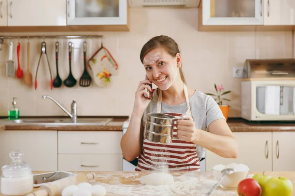 Молодая красивая счастливая женщина просеивает муку с решетом и разговаривает по мобильному телефону на кухне. Готовлю дома. Готовьте еду. . — стоковое фото