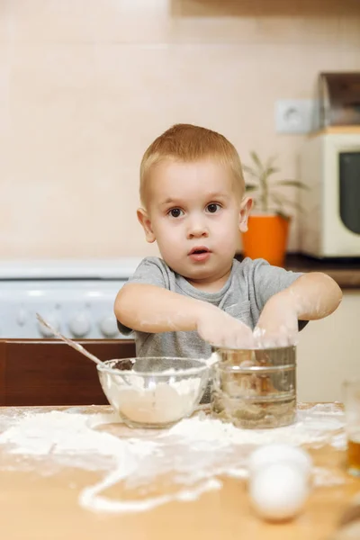 Little kid boy helps mother to cook Christmas ginger biscuit in light kitchen. Happy fair-haired child in gray T-shirt 2-3 years at the table with iron sieve and flour in weekend morning at home. — Stock Photo, Image