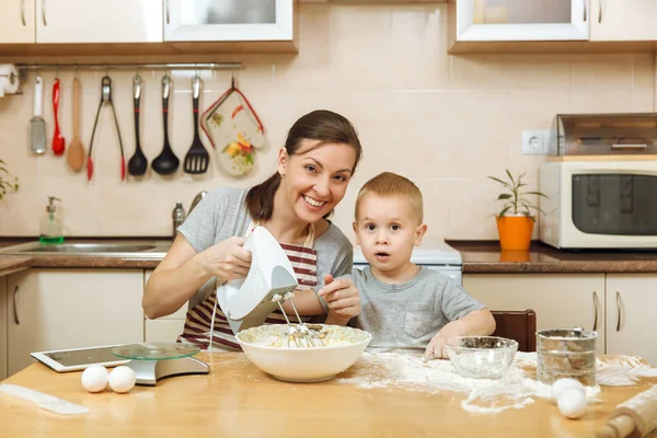 Niño pequeño ayuda a la madre a cocinar galletas de jengibre de Navidad en la cocina ligera con la tableta en la mesa. Feliz familia mamá 30-35 años y el niño 2-3 en la mañana del fin de semana en casa. Concepto de relación —  Fotos de Stock