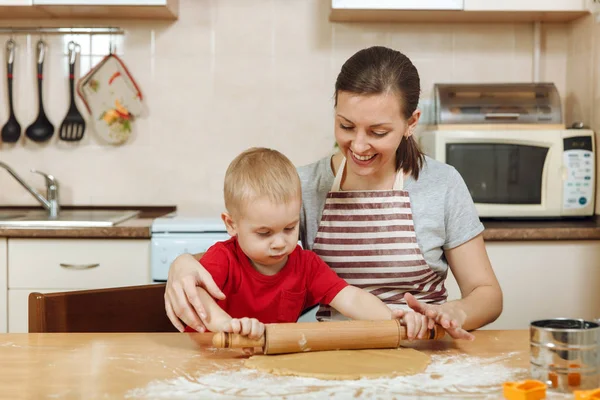 Liten unge pojke hjälper mamma att laga jul ingefära kex i ljus kök. Lycklig familj mamma 30-35 år och barn 2-3 kavla ut degen och skär ut cookies på hem. Relation och kärlek koncept — Stockfoto