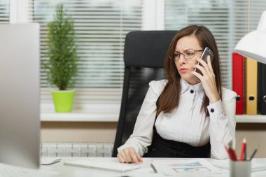 Işık Office belgeleri ile çağdaş bilgisayarda çalışan güzel ciddi iş kadın takım elbise ve masa başında oturan gözlük cep telefonu üzerinde sorunları çözme bir kenara seyir konuşurken