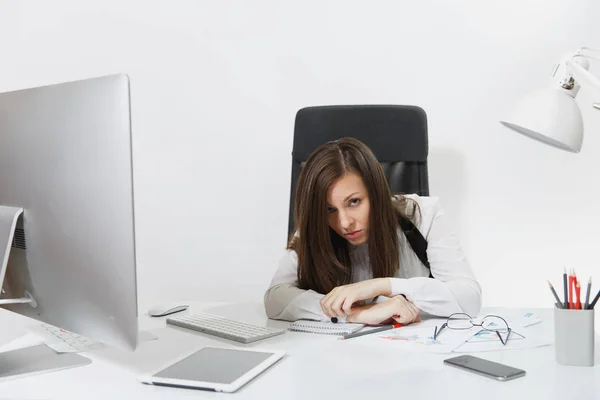 Красивая уставшая смущенная и напряженная деловая женщина в костюме и очках сидит за столом, работает за современным компьютером с документами и монитором в светлом офисе на белом фоне — стоковое фото
