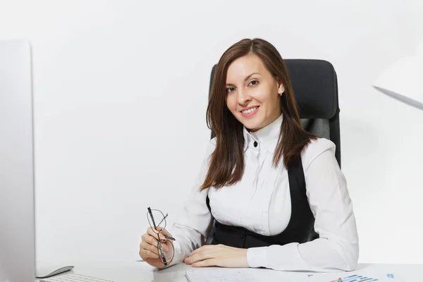 Beautiful smiling brown-hair business woman in suit and glasses sitting at the desk, working at computer with modern monitor with documents in light office, looking at the camera on white background Stock Image