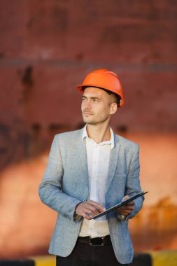 Yakışıklı tıraşsız başarılı iş adamı gri takım elbise, tablet, liman kargo paslı gemi arka plan ayakta tutan koruyucu inşaat turuncu kask. Erkek ile güneşli günde becerikli alet